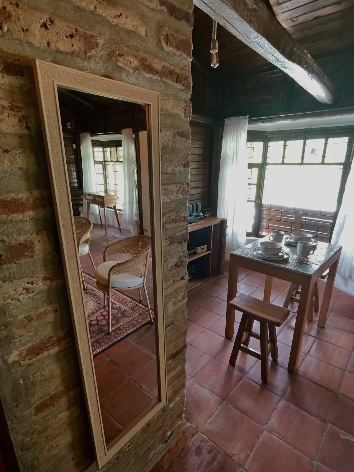 Interior de la cabaña de la quinta: paredes de ladrillo visto, piso de baldosa, espejo de pie y comedor de madera junto a la ventana