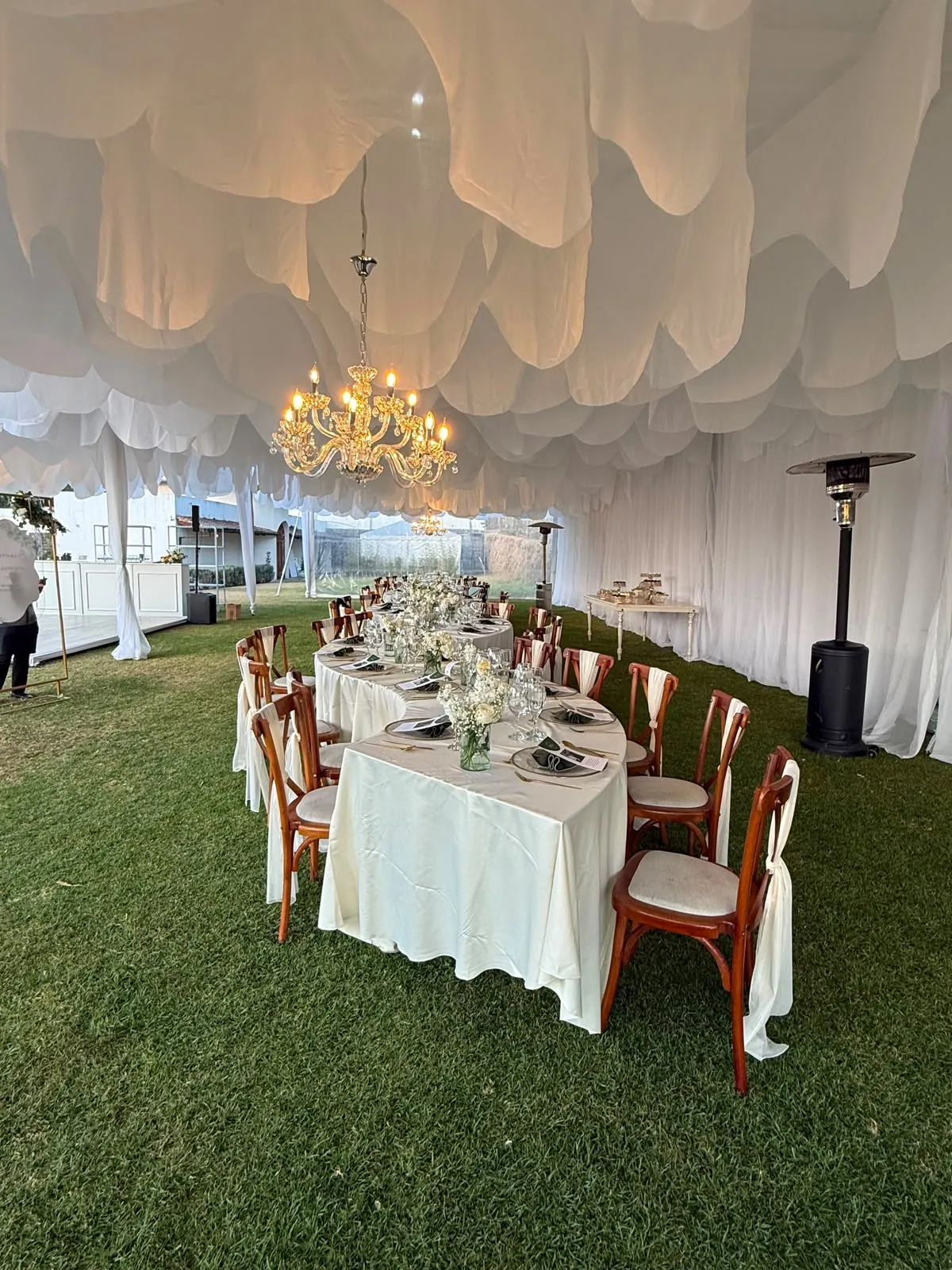 Carpa con techo de telas blancas y candelabro de cristal sobre una mesa larga montada sobre el césped