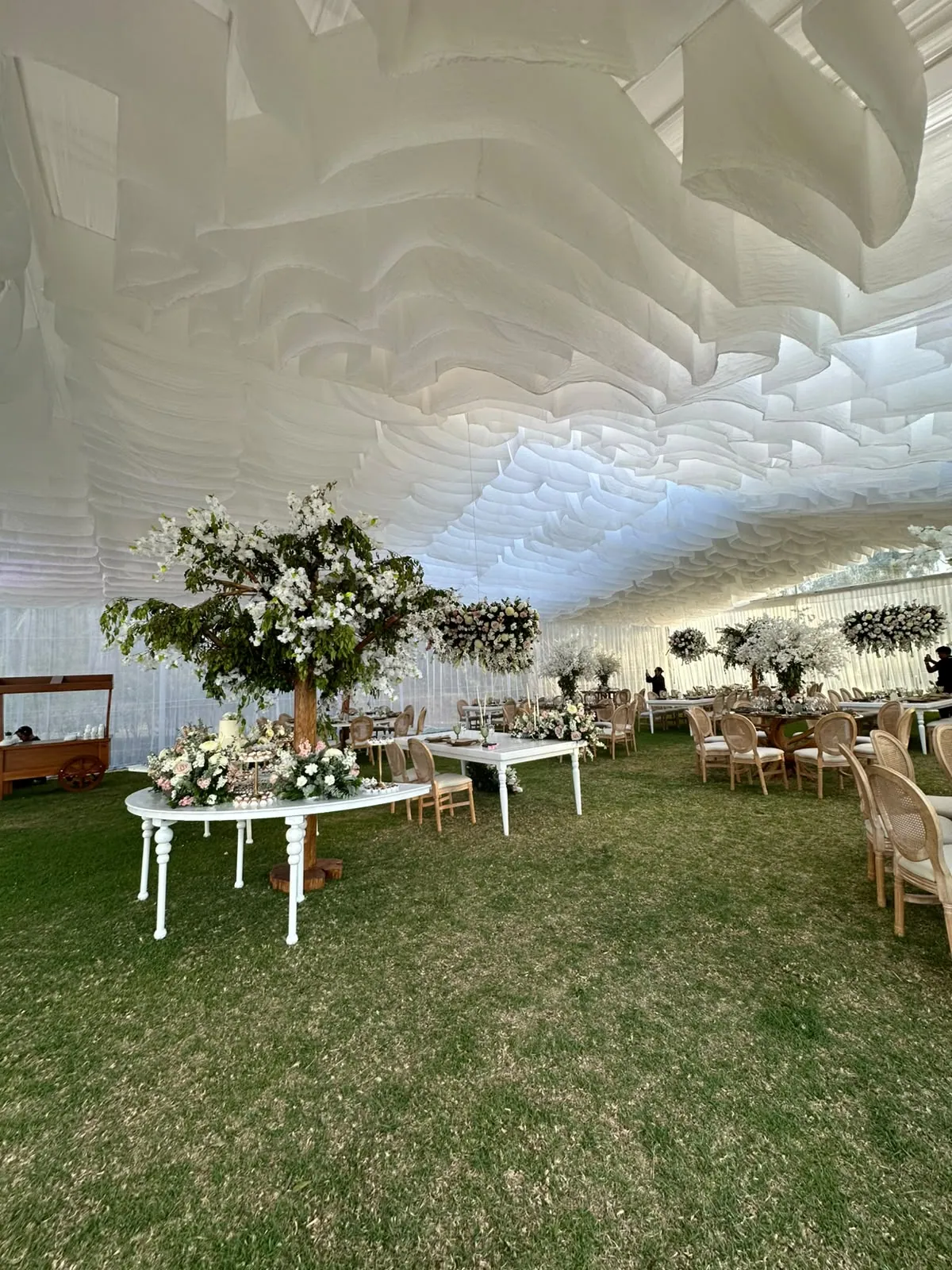Vista general de la carpa de la Quinta Las Tres Marías montada sobre el césped, con mesas vestidas en blanco, centros florales altos y un carrito de madera para catering