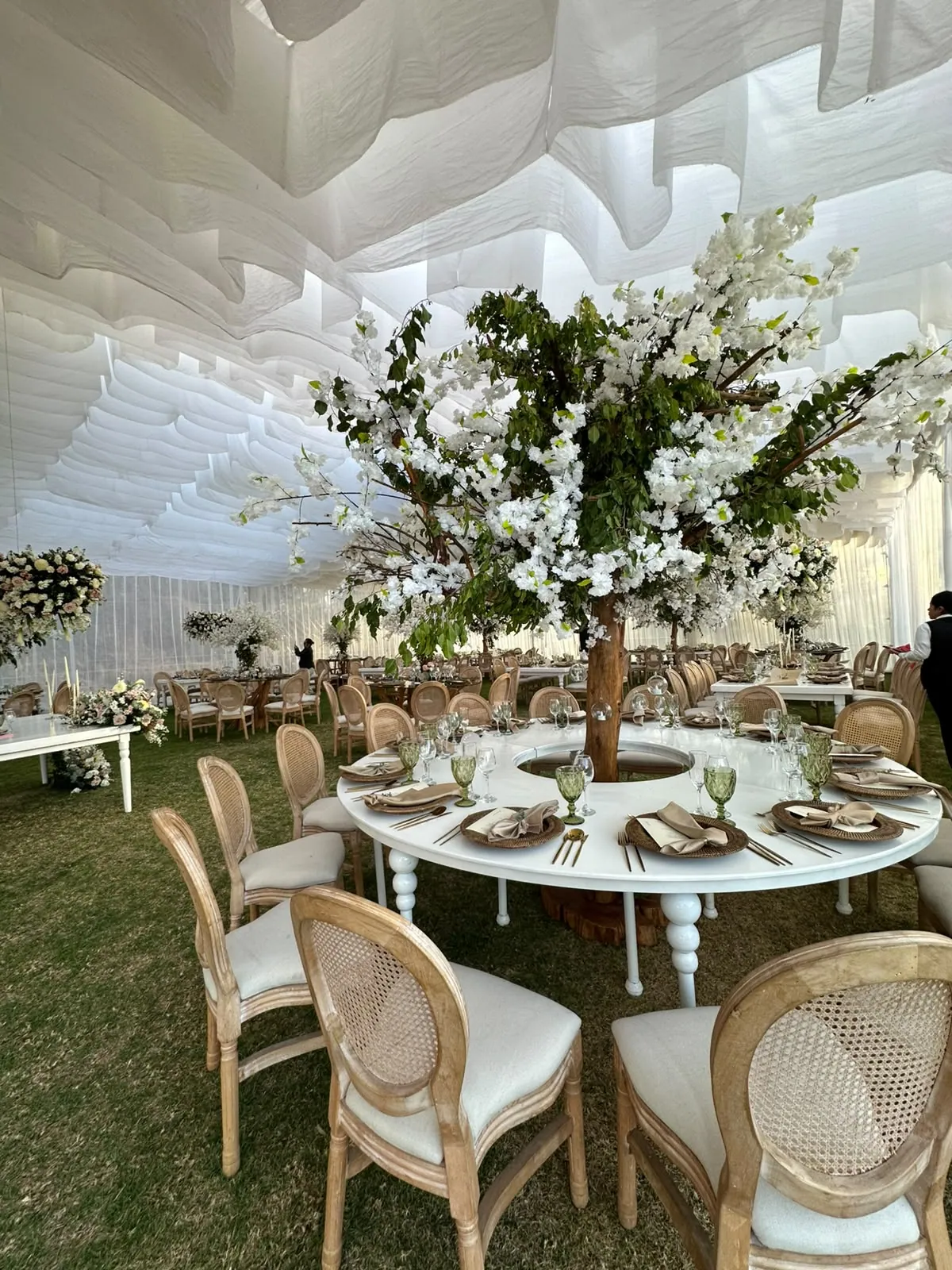 Mesa redonda bajo la carpa de la Quinta Las Tres Marías, vestida en blanco con sillas de ratán, vajilla y un árbol de flores blancas como centro de mesa