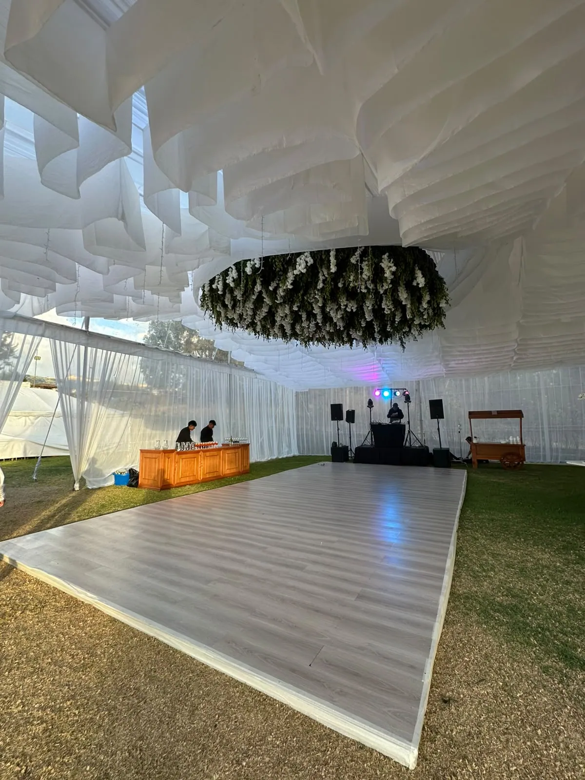 Pista de baile bajo la carpa de la Quinta Las Tres Marías, con barra de bebidas, cabina de DJ con luces de colores y un arreglo floral colgante en el techo